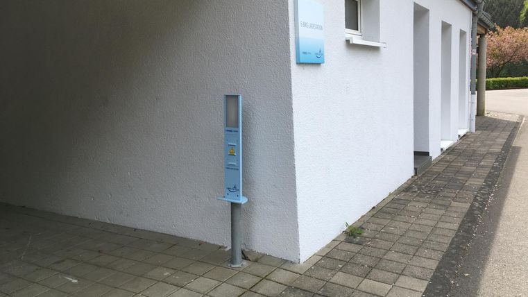 Ein weißes Gebäude mit einem Gehweg daneben. An der Wand befindet sich ein Informationsständer.