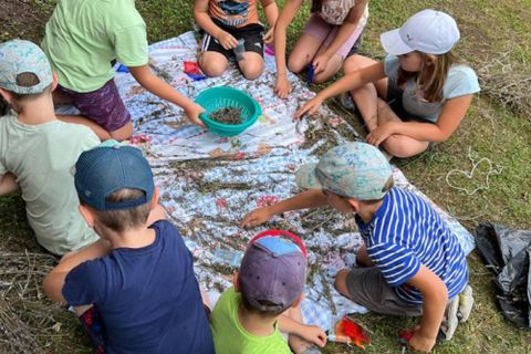 Eine Gruppe von Kindern sitzt auf einem Tuch und spielt mit verschiedenen Materialien. Sie verwenden eine Schüssel und wirken konzentriert und glücklich.