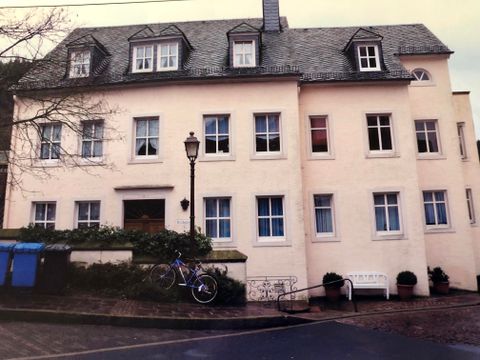A multi-story, bright building with many windows. In front of the building, there is a bicycle and a bench, surrounded by some plants.