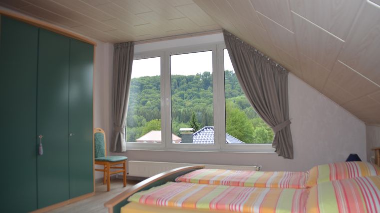 A bright bedroom with a double bed and colorful bedding. The view from the window shows a green hilly landscape.