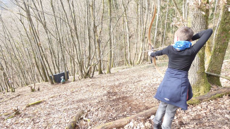 Een jongen schiet met een boog in een bos. De omgeving is gekenmerkt door bomen en bladeren.