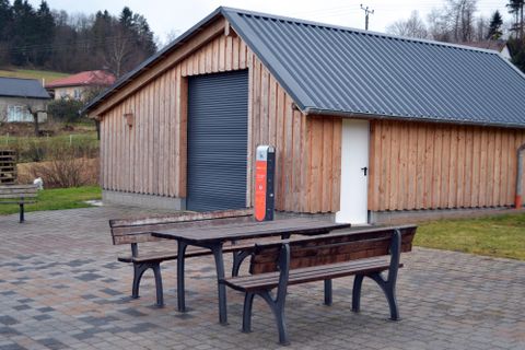 Ein Holzgebäude mit einem großen Rollladen und einem weißen Tor. Vor dem Gebäude stehen zwei Holztische und Bänke auf einem gepflasterten Bereich.