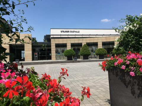 Ein modernes Gebäude mit großen Fenstern und einem klaren blauen Himmel. Bunte Blumen in Töpfen rahmen den Platz ein.