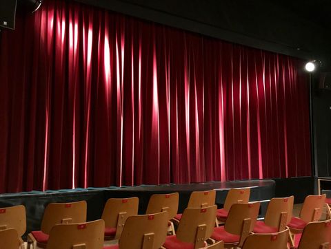 Ein Theater mit roten Vorhängen auf der Bühne. Vor der Bühne stehen mehrere Stühle in Reihen.