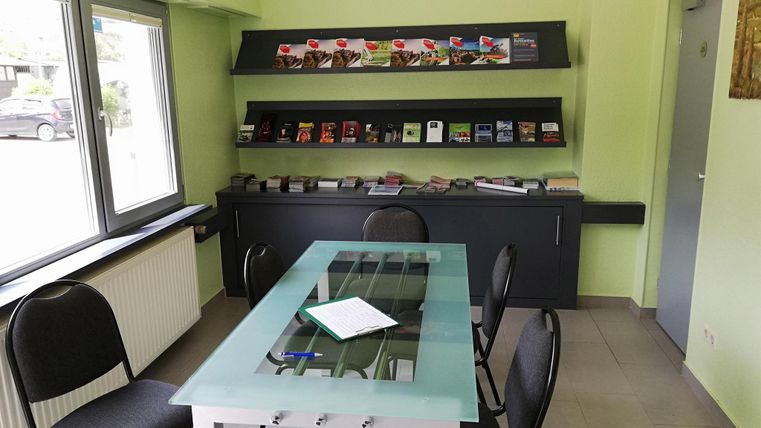 Een eenvoudige vergaderzaal met een glazen tafel en zwarte stoelen. Op de achtergrond staat een rek met verschillende brochures.