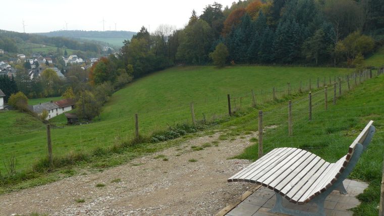 Een bank met uitzicht op Mettendorf, omgeven door groene velden en bomen.