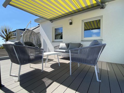 Ein gemütlicher Außenbereich mit zwei modernen Sesseln und einem Tisch. Über dem Bereich spannt ein gestreifter Sonnenschirm und im Hintergrund ist eine Hängesessel zu sehen.