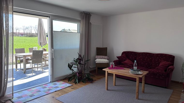 Ein gemütliches Wohnzimmer mit einem roten Sofa und einem Holztisch. Große Fenster bieten einen Blick auf die Terrasse und den Garten.