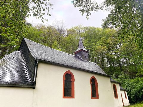 Eine kleine Kirche mit einem spitzen Turm, umgeben von grünen Bäumen. Der Himmel ist bewölkt und die Fassade der Kirche hat rot eingerahmte Fenster.