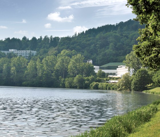 Seeufer des Stausees Bitburg mit grünen Bäumen, einem Gebäude im Hintergrund und klarem Himmel., © TI Bitburger Land