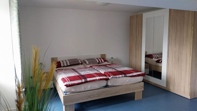 Ein modernes Schlafzimmer mit einem großen Bett und karierter Bettwäsche. Ein schmaler Kleiderschrank und grüne Pflanzen sorgen für eine gemütliche Atmosphäre.