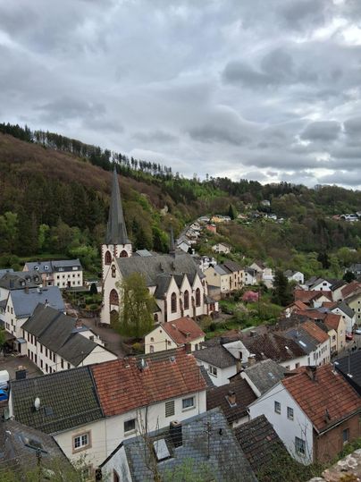 Une vue pittoresque du village avec une église au premier plan et des collines douces en arrière-plan. Le ciel est nuageux et les maisons sont de différentes couleurs.