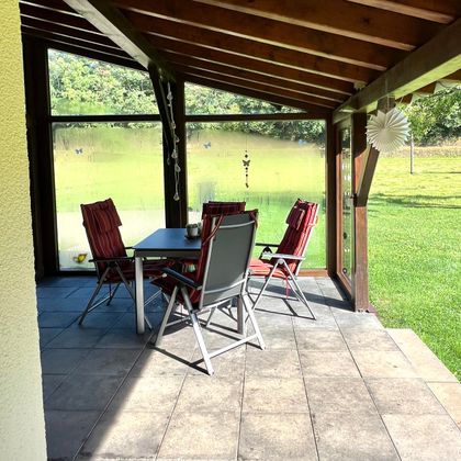 Eine gemütliche Terrasse mit einem Esstisch und vier Stühlen. Im Hintergrund ist eine grüne Wiese sichtbar.