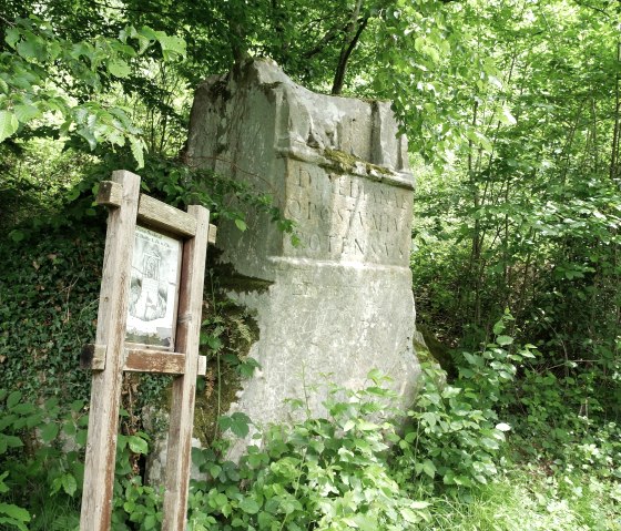 Ein Steinmonument im Wald, umgeben von &uuml;ppigem Gr&uuml;n. Daneben steht eine h&ouml;lzerne Informationstafel., &copy; Elke Wagner, Felsenland S&uuml;deifel Tourismus GmbH