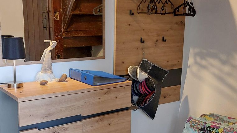 Een moderne entree met houten kasten en een spiegel. Aan de muur hangen kledinghaken en er is een kleurrijke, gewatteerde zitgelegenheid.