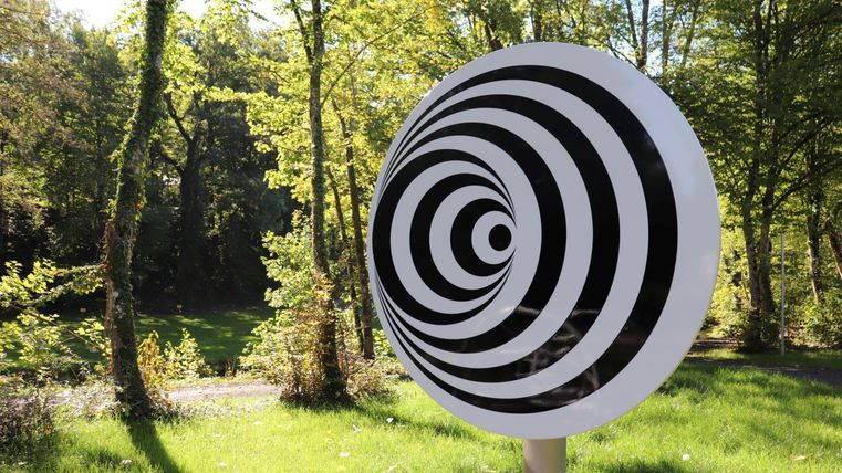 Ein großes, kreisförmiges Kunstwerk mit schwarzen und weißen Spiralen steht auf einer Wiese. Im Hintergrund sind grüne Bäume und ein ruhiger Weg zu sehen.