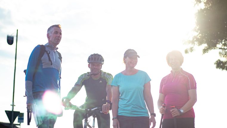 Eine Gruppe von vier aktiven Personen steht im Freien, eine Person sitzt auf einem Fahrrad. Die Sonne scheint hell im Hintergrund, wodurch eine angenehme Atmosphäre entsteht.
