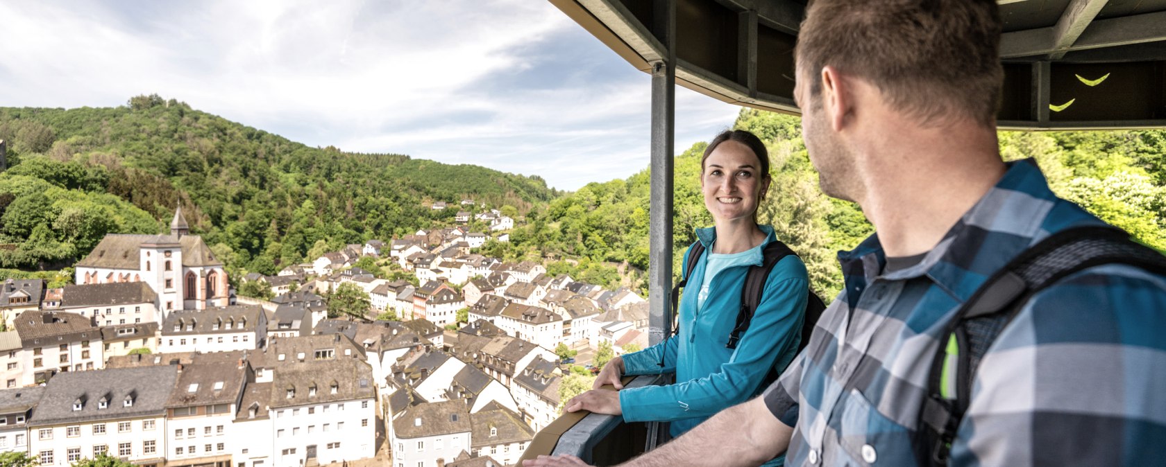 Zwei Personen stehen auf einem Aussichtspunkt und blicken auf eine malerische Stadt mit Kirche und grünen Hügeln im Hintergrund., © Eifel Tourismus GmbH, Dominik Ketz