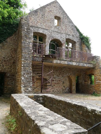 Eine alte Steinruine mit einem oberen Balkon und einer Wendeltreppe. Rundherum sind Pflanzen und Bäume zu sehen.