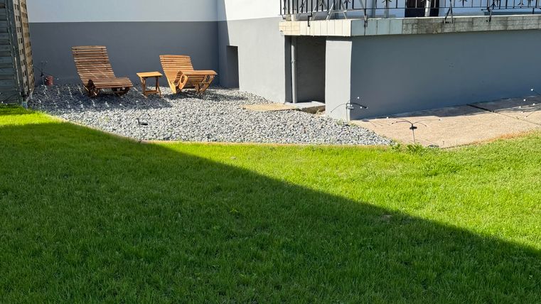 Ein modernes Haus mit einem gepflegten Garten. Zwei Liegen stehen auf einem Kiesbereich neben der Rasenfläche.