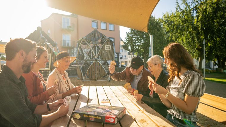 Eine Gruppe von sechs Personen sitzt an einem Tisch und spielt ein Kartenspiel. Im Hintergrund sind Spielgeräte und ein sonniger Park zu sehen.