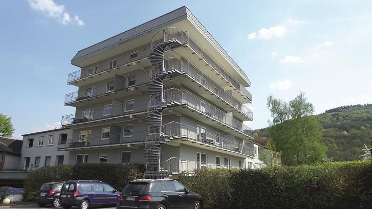 Ein modernes Mehrfamilienhaus mit vier Etagen und einem Stahlaußensteg. Im Vordergrund stehen mehrere Autos auf einem Parkplatz.