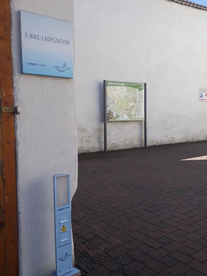 Eine weiße Wand mit einem Informationsschild und einer Karteninformation. Der Boden ist gepflastert und der Himmel ist blau.