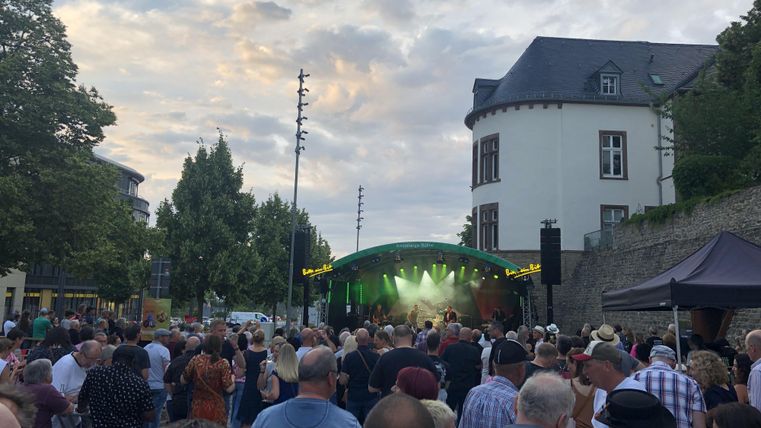 Eine Menschenmenge bei einem Open-Air-Konzert vor einer Bühne. Im Hintergrund sind Bäume und Gebäude sichtbar, während der Himmel teilweise bewölkt ist.