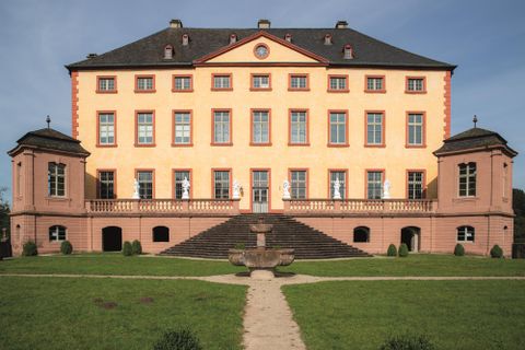 Ein beeindruckendes Schloss mit einer orangefarbenen Fassade und großen Fenstern. Es liegt umgeben von einem gepflegten Garten und bietet eine majestätische Aussicht.
