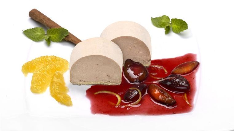 Een elegant dessert van een romige bereiding, geserveerd met een fruitige saus en garnering van stukjes sinaasappel en muntbladeren. Een kaneelstokje maakt de aantrekkelijke presentatie compleet.
