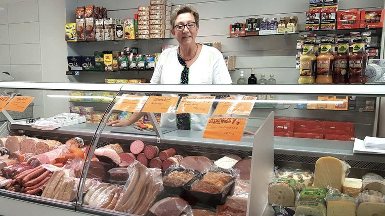 A friendly saleswoman stands behind a meat and cheese counter. The display offers a wide selection of sausages and cheese varieties.