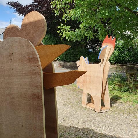 Zwei Holzskulpturen von Tieren stehen im Freien. Eine Figur zeigt einen Hund mit einem leuchtend roten Schwanz und eine andere Figur hat die Form eines Huhns.