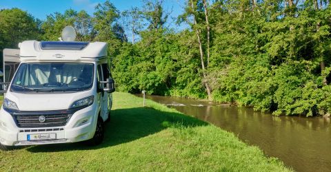 Een camper staat aan de oever van een rustige rivier, omringd door groene bomen. De lucht is helder en zonnig.