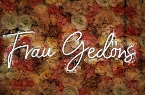 Ein Schriftzug "Frau Gedöns" vor einem Hintergrund aus bunten Blumen. Die Blüten sind in zarten Rosa- und Beigetönen gehalten.
