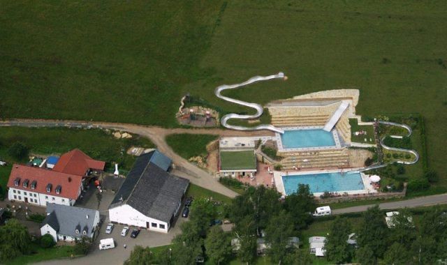 Eine Luftaufnahme von einem Freizeitbereich mit mehreren Pools und Rutschen. Umgeben von grünen Wiesen und einer ruhigen Landschaft.