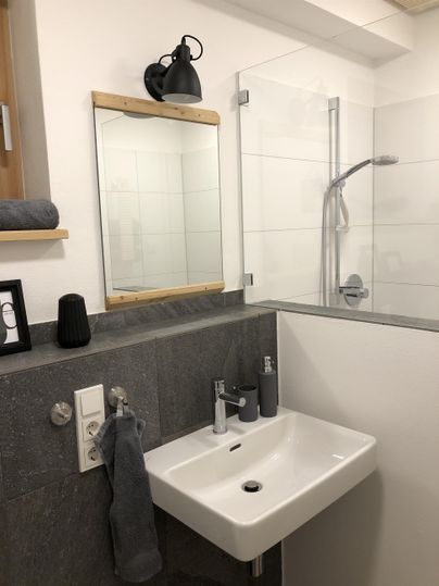 Ein modernes Badezimmer mit einem eleganten Waschbecken und einem großen Spiegel. Die Dusche ist mit Glaswänden abgetrennt und die Wände sind hell und zeitgemäß gestaltet.