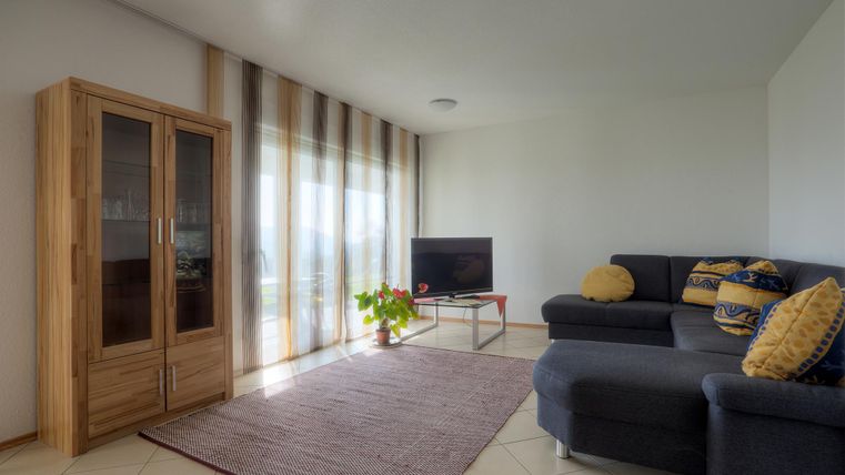 Ein modernes Wohnzimmer mit einem grauen Sofa und einem Teelichthalter auf einem kleinen Tisch. Große Fenster lassen viel Licht herein, und es gibt eine Holzvitrine sowie einen Teppich.