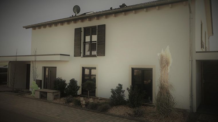 Ein modernes Haus mit weißen Wänden und dunklen Fensterläden. Es gibt Pflanzen im Vordergrund und eine Satellitenschüssel auf dem Dach.