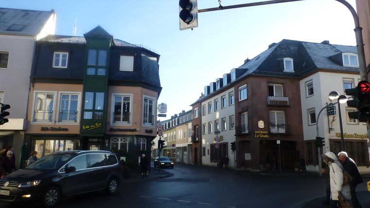 Eine Straßenszene mit mehreren Gebäuden und einem Ampelmast. Auf der Kreuzung warten Autos und Fußgänger auf das Lichtsignal.