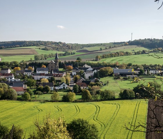 Panoramic view of the village of Alsdorf with a church in the center, surrounded by green fields and hills under a blue sky., © Tourist-Info Bitburger Land