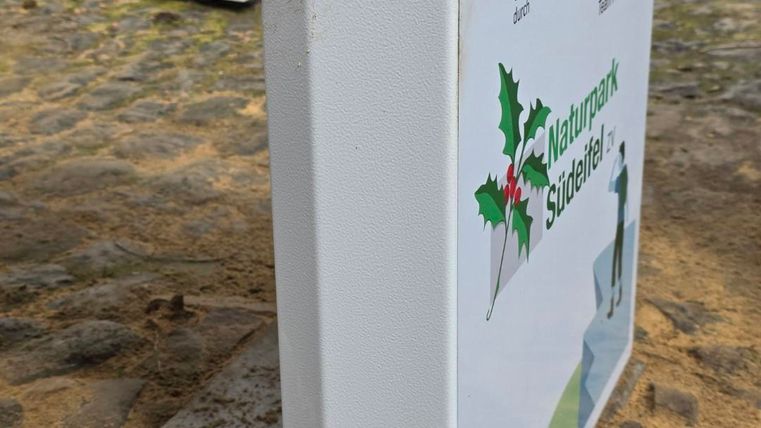 Ein Informationsschild über die Natur in einem parkähnlichen Gebiet. Das Schild hat eine klare, weiße Oberfläche mit grünen und bunten Symbolen.