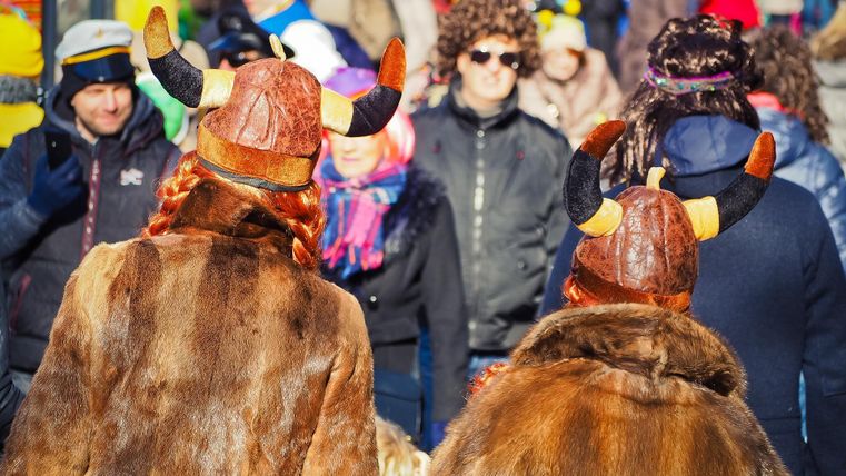 Zwei Personen in Wikingerkostümen mit Hörnerhelmen stehen im Mittelpunkt einer fröhlichen Menschenmenge. Es ist eine lebhafte Veranstaltung, die mit bunten Kleidern und Spaß gefüllt ist.