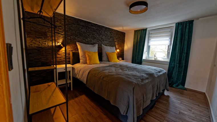 Une chambre confortable avec un grand lit, des coussins moelleux et des rideaux verts. Sur le mur, il y a un design rustique et une lampe de chevet claire.