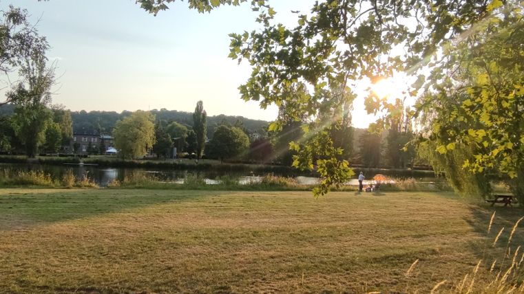 Eine ruhige Wiese am Fluss mit Sonnenstrahlen im Hintergrund. Bäume umgeben die Szene, und im Vordergrund sitzen zwei Personen auf einer Bank.