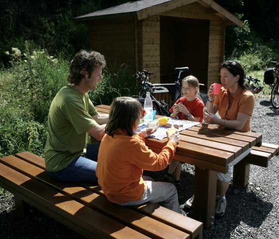 Eine Familie sitzt an einem Holztisch im Freien und macht eine Pause. Im Hintergrund stehen Fahrräder und eine Holzhütte., © Eifel Tourismus GmbH/intention