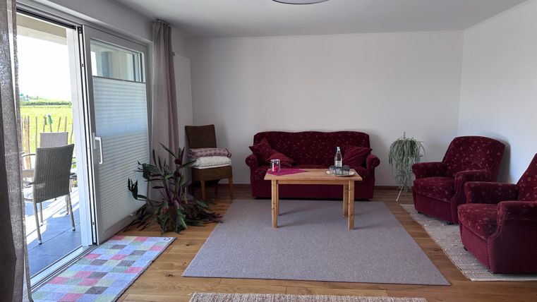 Ein gemütlicher Wohnraum mit roten Sofas und einem Holztisch. Große Fenster lassen viel Licht herein und bieten einen Blick nach draußen.