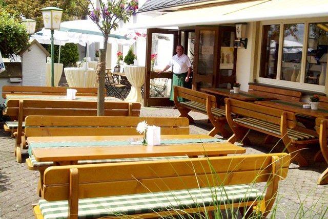 Un espace extérieur accueillant d'un restaurant avec des tables en bois et des bancs. À l'arrière-plan, on voit une personne entrer dans le restaurant.