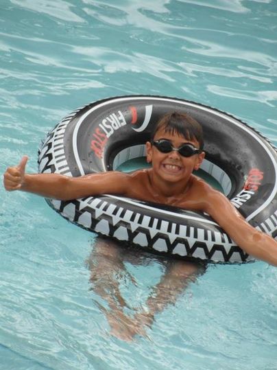 Ein fröhlicher Junge schwimmt in einem Pool und trägt eine Schwimmbrille. Er sitzt in einem Gummireifen und zeigt Daumen nach oben.