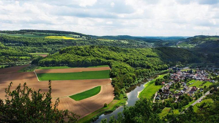 A picturesque landscape with gentle hills, fields, and a river flowing through the valley. In the background, forests and a small village are visible.