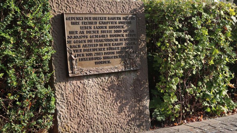 Eine Gedenktafel aus Stein mit einer Inschrift zur Erinnerung an historische Ereignisse. Sie steht in einem bepflanzten Bereich neben einem Gehweg.